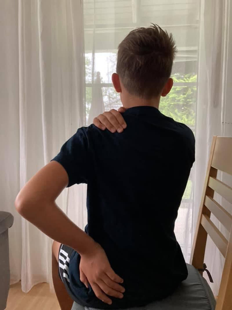 Junger Mensch sitzt mit dem Rücken zur Kamera und fasst mit einer Hand an die Schulter, mit der anderen an den unteren Rücken